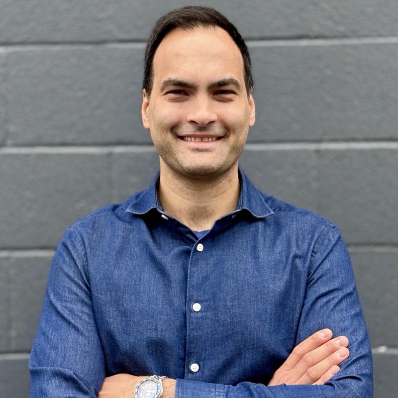 Birkan Icacan, smiling, arms crossed, in a blue shirt against a grey wall